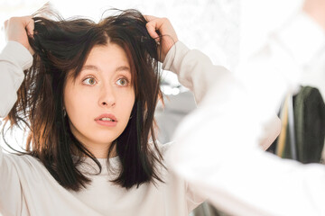 A young woman stands in front of a mirror and looks in surprise at the condition of her hair, seasonal problem of hair loss, oily, dry, unkempt hair