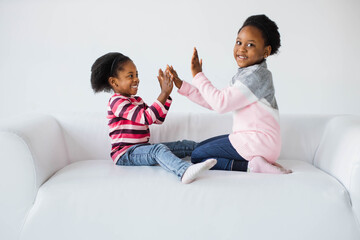 Two pretty african sisters sitting on white sofa and playing together. Cute female children in casual outfit smiling sincerely while taking fun during leisure time.