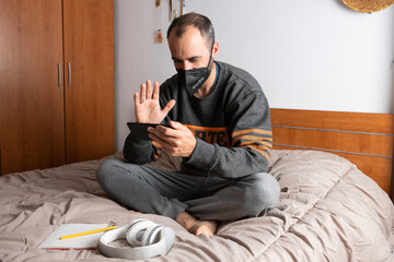 Man with headphones, mobile phone and face mask, sitting on his bed and confined to his room