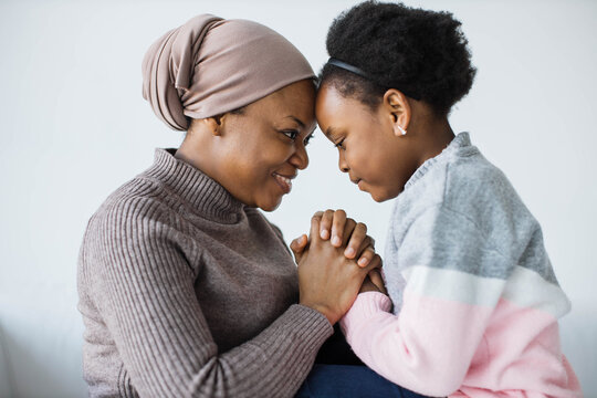 African american mother in headscarf and her little daughter sitting face to face, holding hands and keeping eyes closed. Studio with white background.