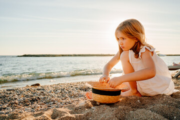 girl in white dress sits on seashore and collects pebbles and twigs in straw hat