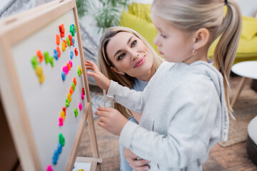 Obraz premium Smiling mother looking at daughter near easel with colorful magnets at home.