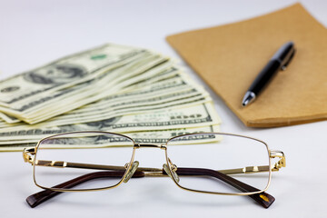 Gold-rimmed eyeglasses lie in the foreground, and a stack of American dollars, a notepad, and a pen are in the background. 