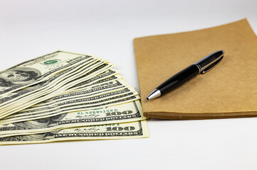A stack of American 100 dollar bills, next to it is a kraft notebook and a premium pen.