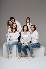Delighted cute young girls, their moms and grandmother smiling, hugging while sitting on white cubes on gray background wearing jeans and white shirts. Lovely women group