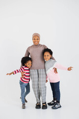 Fototapeta premium Full length portrait of happy african muslim woman in hijab embracing her smiling daughters while standing over white background. Family leisure and playful mood concept.