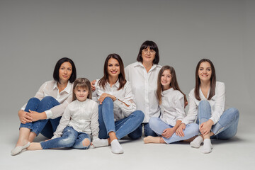 Delighted cute young girls, their moms and grandmother smiling, sitting on gray background wearing jeans and white shirts. Lovely women group