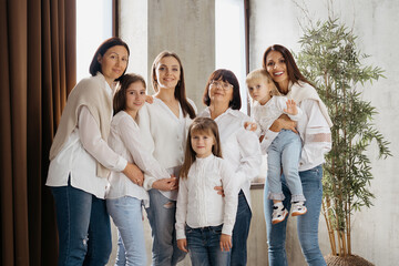 Delighted cute young girls, their moms and grandmother smiling, hugging while standing at home in the living room wearing jeans and white shirts. Lovely women group