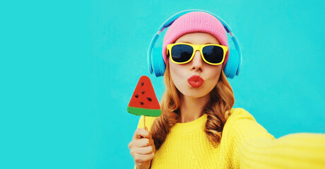 Summer fresh colorful portrait of young woman taking selfie in headphones listening to music with fruit juicy lollipop or ice cream shaped slice of watermelon on blue background