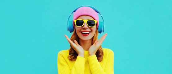 Portrait of happy surprised smiling young woman model with headphones listening to music wearing colorful yellow sweater on blue background