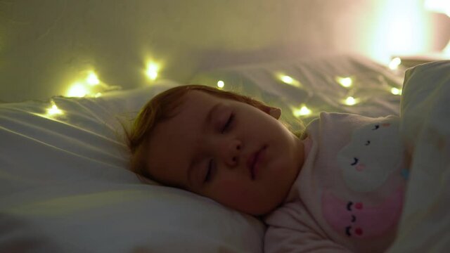 Cute Chuby Little Toddler Baby Girl 1-2 Years Old Sleeping In Pink Pajama On White Bed In Dark Bedroom. Child Have Relax Rest Time At Night Garland on Pillow. Childhood, Christmas, Parenthhood Concept