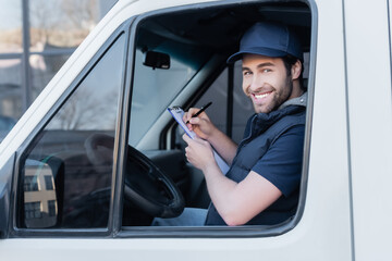 Young courier smiling at camera while writing on clipboard in car.
