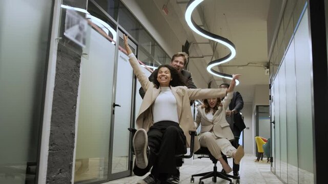 Millennial multiracial team people having fun riding on chairs in office room, excited diverse employees laughing enjoying funny activity at work break, creative friendly workers play game together