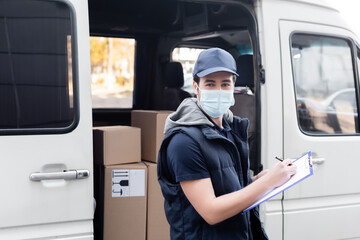 Delivery man in medical mask writing on clipboard near carton boxes in car outdoors.