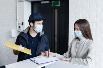 Businesswoman in medical mask writing on clipboard near courier with parcel in office.