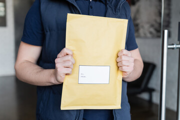 Cropped view of parcel with sticker in hands of blurred courier in hallway.
