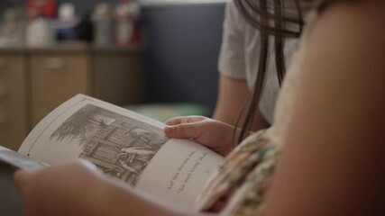Close up of young girl reading book in classroom with teacher sitting near - Powered by Adobe