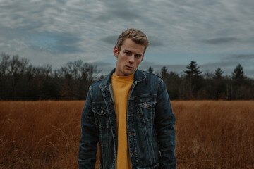 Man with vintage style wearing a jean jacket standing in an open field looking at camera sad and depressed 