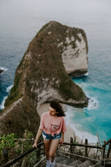 A young travelling and backpacking girl visits Kelingking Beach in Nusa Penida, Indonesia. Overlooking crystal clear turquoise water, waves, blue ocean and sky on a sunny day.