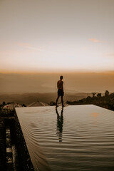 A young male traveller and blogger is walking on the edge of infinity pool at a luxury jungle resort in Bali, Indonesia at sunset.