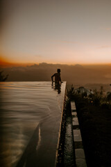 A young male traveller and blogger is walking on the edge of infinity pool at a luxury jungle resort in Bali, Indonesia at sunset.