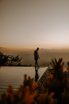 A young male traveller and blogger is walking on the edge of infinity pool at a luxury jungle resort in Bali, Indonesia at sunset.
