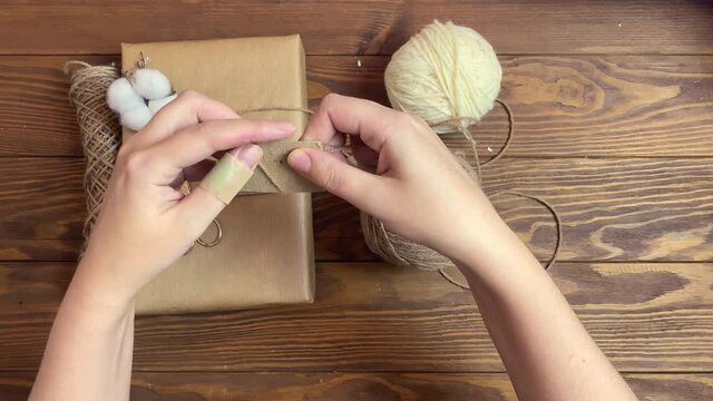 Women's hands wind brown yarn on piece of cardboard. Box in craft wrapper, twine, cotton and scissors on wooden table. Handmade crafting decorations of gift for Christmas or Birthday. Festive mood.