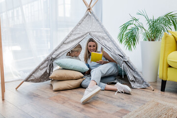 Mother reading book near child in teepee at home.