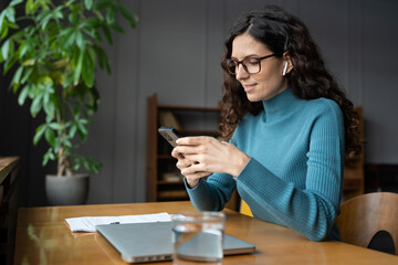 Young businesswoman use smartphone online apps for business social media management and ads. Female distance manager worker checking email messages on mobile phone sitting at desk with closed laptop
