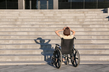 Stressed young woman in wheelchair near stairs outdoors