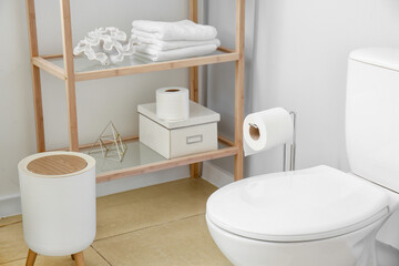 Toilet bowl, holder with roll of paper and shelf unit near light wall in restroom interior
