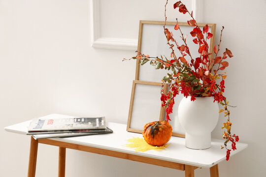 Stylish vase with branches, blank frames, magazines and pumpkin on table near white wall
