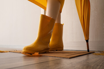 Woman in yellow raincoat and gumboots on floor mat near light wall