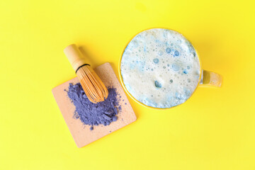 Fototapeta na wymiar Cup of blue matcha latte, powder and chasen on yellow background