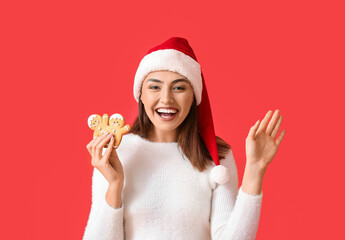 Pretty young woman in Santa hat with gingerbread cookies on red background