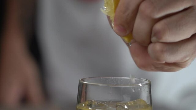 Hand squeezing lemon into freshly made cocktail in tall clear glass with ice cubes close up