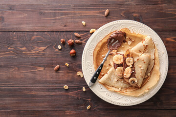 Plate of tasty thin pancakes with chocolate paste, cut banana and nuts on wooden background