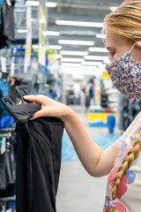 New normal retail shopping. Teenage blond girl wearing face mask choosing sports clothing at sport warehouse retail shop. - obrazy, fototapety, plakaty