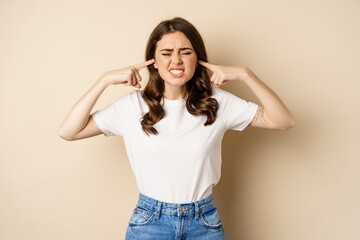 Annoyed young woman shut ears from loud noise, feeling discomfort and complaining, standing against...