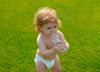 Baby drinks mineral water from bottle. Child resting in grass park.