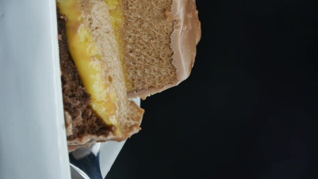 a steel dessert spoon cuts a chocolate cake with mango filling and biscuit on a white ceramic plate and a black background. dollar shot dessert. Vertical screen, copy space