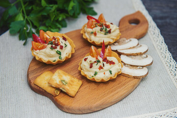 on a table on a gray background on a cutting board are three tortelets with hummus with slices of tomato and spicy seasoning, slices of mushrooms and crackers lie side by side in the background green 