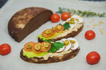 on the table on a light background lies half a loaf of black bread, next to it are two sandwiches with hummus salad, tomatoes, olives and an egg. Cherry tomatoes are also nearby and a green twig lies 