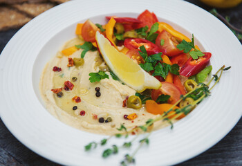 in a white plate hummus with a slice of lemon chopped hot pepper slices of tomato and spicy seasoning with sprigs of greenery on the edge