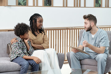 Confident doctor in uniform consulting African female with her son sitting on couch in front of him...