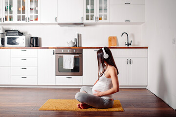 Pregnant doing workout on orange fitness mat. Pragnant yoga at home in the kitchen. Pregnant in white headphones.