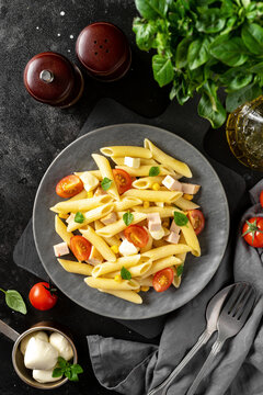 Pasta salad with ham, mozzarella, corn, tomatoes and olive oil in a ceramic plate on a dark culinary background. Traditional Italian dish penne rigate with cheese and vegetables top view