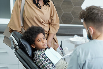 Cute African boy looking at dentist while sitting in armchair against his mother supporting him...