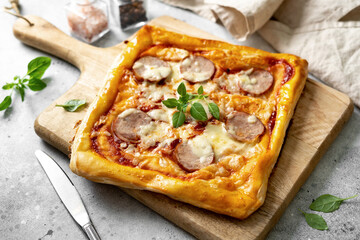 Homemade puff pastry pizza with salami, mozzarella, tomatoes and basil on the kitchen table. Delicious layered pie with sausage and cheese on a light culinary background 