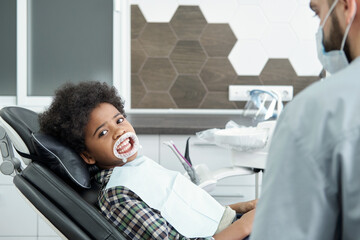 Little boy with medical retractor on mouth looking at you while sitting in armchair in dental...